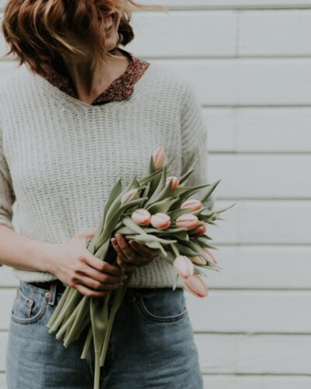 A woman in a cozy sweater holding a bouquet of pink tulips, smiling with her hair slightly tousled. Soft, natural tones create a warm and inviting feel.